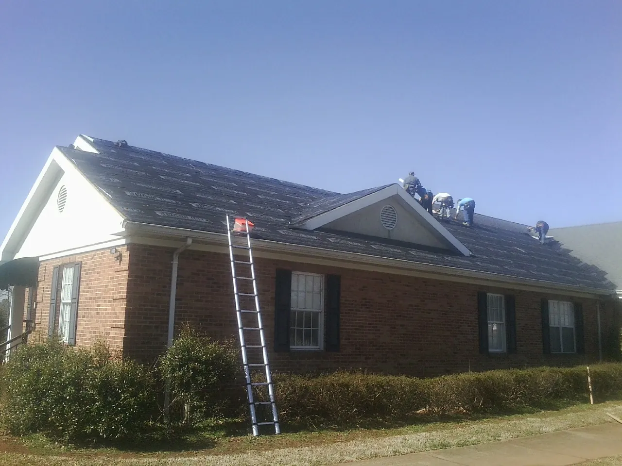 Crew tearing shingles off a brick ranch roof with a ladder set — Emergency Roof Repair work by our crew in Cohasset