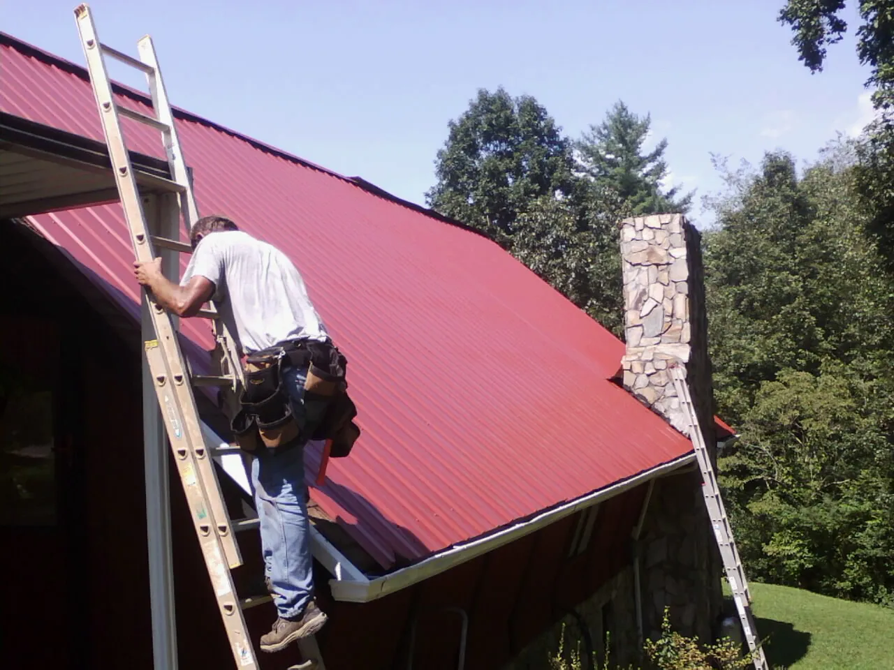 Roofer on a ladder beside a red metal roof and stone chimney — Soffit & Fascia Repair by Roofing Brothers in Cohasset