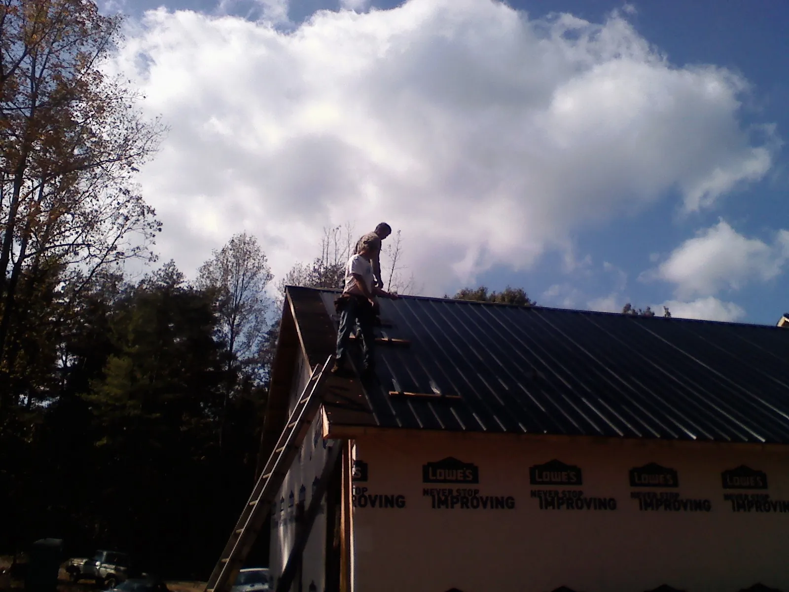 Roofer fastening dark metal panels high against a cloudy sky, from a Roof Inspection job in Cohasset